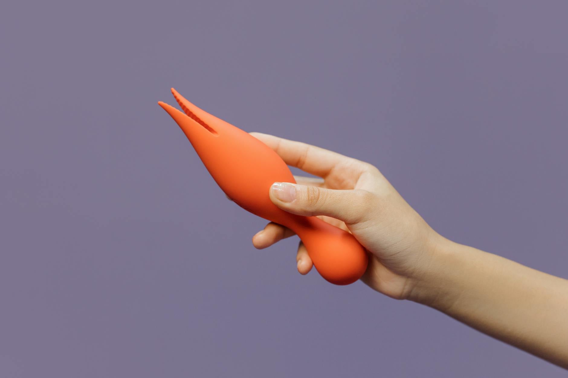 A hand holding a yellow and blue personal vibrator against a minimalist purple backdrop